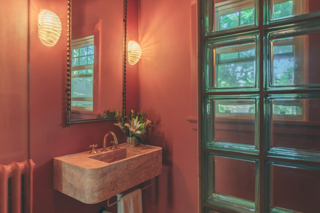 Dramatic powder room with coral walls pink travertine sink and green glass block window Denver. Interior Design by Tenlie Mourning